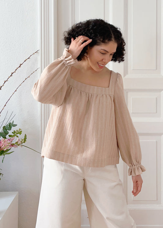 Woman wearing the Alba blouse in light brown organic cotton standing against a white paneled wall.
