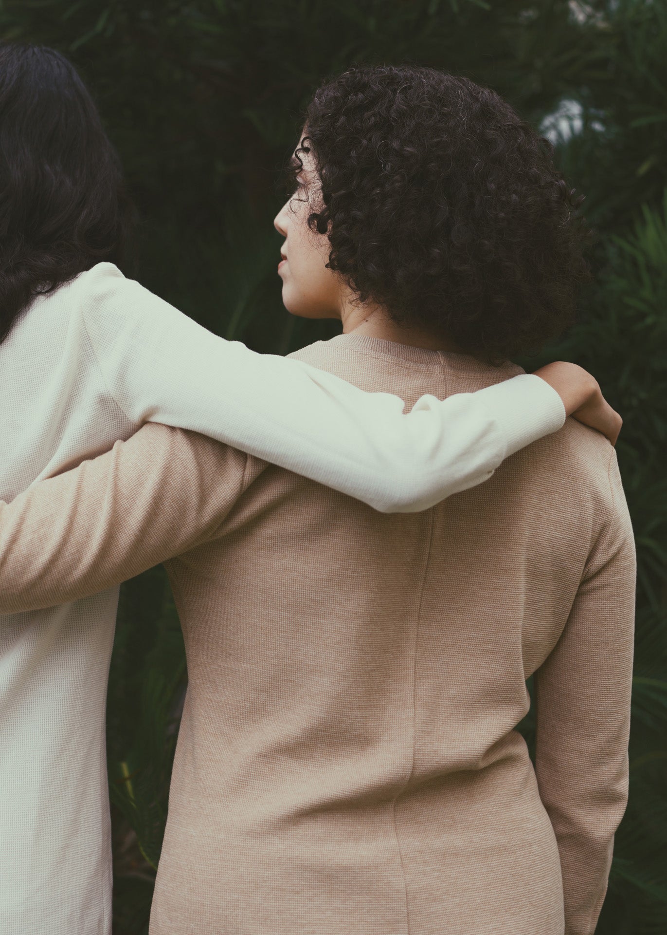 Two women standing close together with one person's arm around the other, against a background with plants. They are both wearing organic cotton dresses with long sleeves.