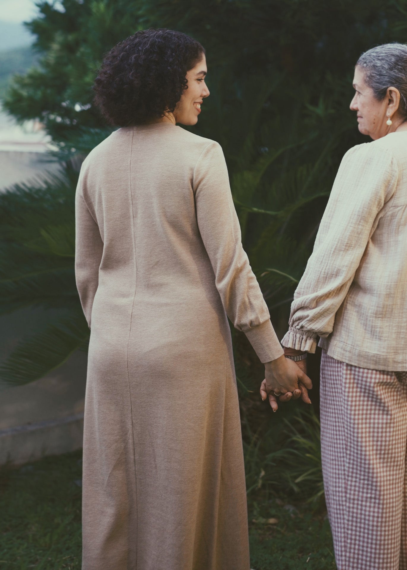Two women holding hands outdoors with a natural background. The woman on the left is wearing a long brown dress with a center back seam.
