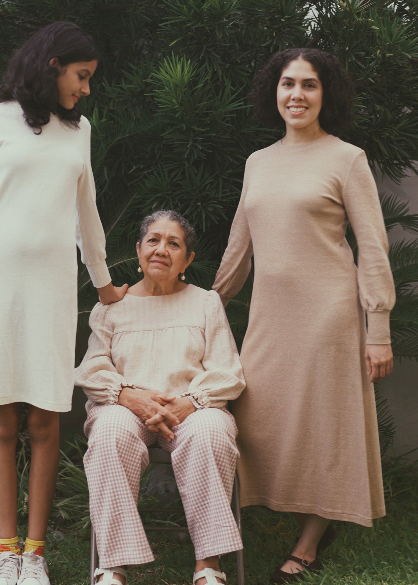 Three women in a garden setting, with one sitting and the other two standing. The one on the right is wearing a midi dress in light brown organic cotton.