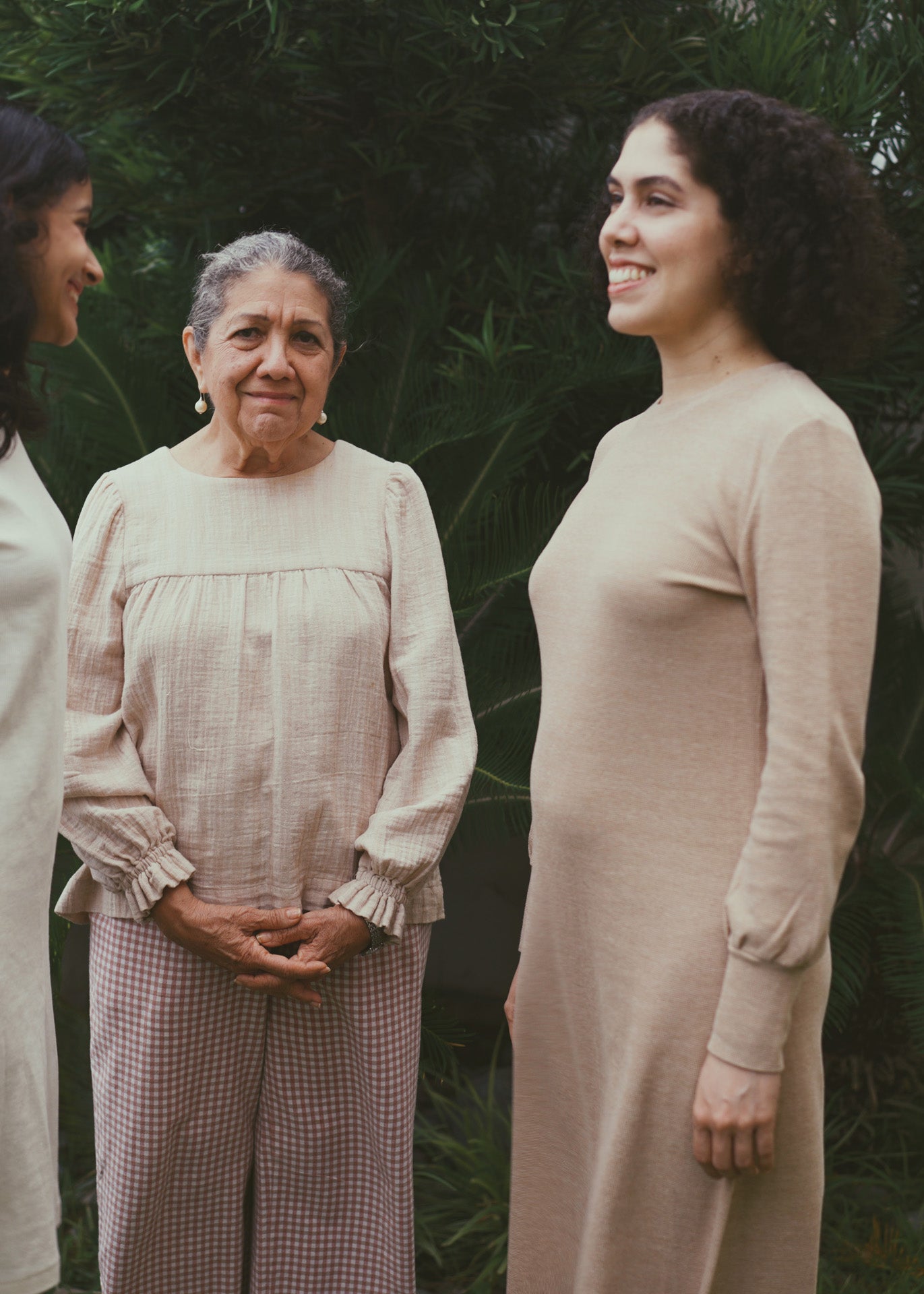 Three women standing together outdoors with a natural background. They are smiling and wearing organic cotton clothing.