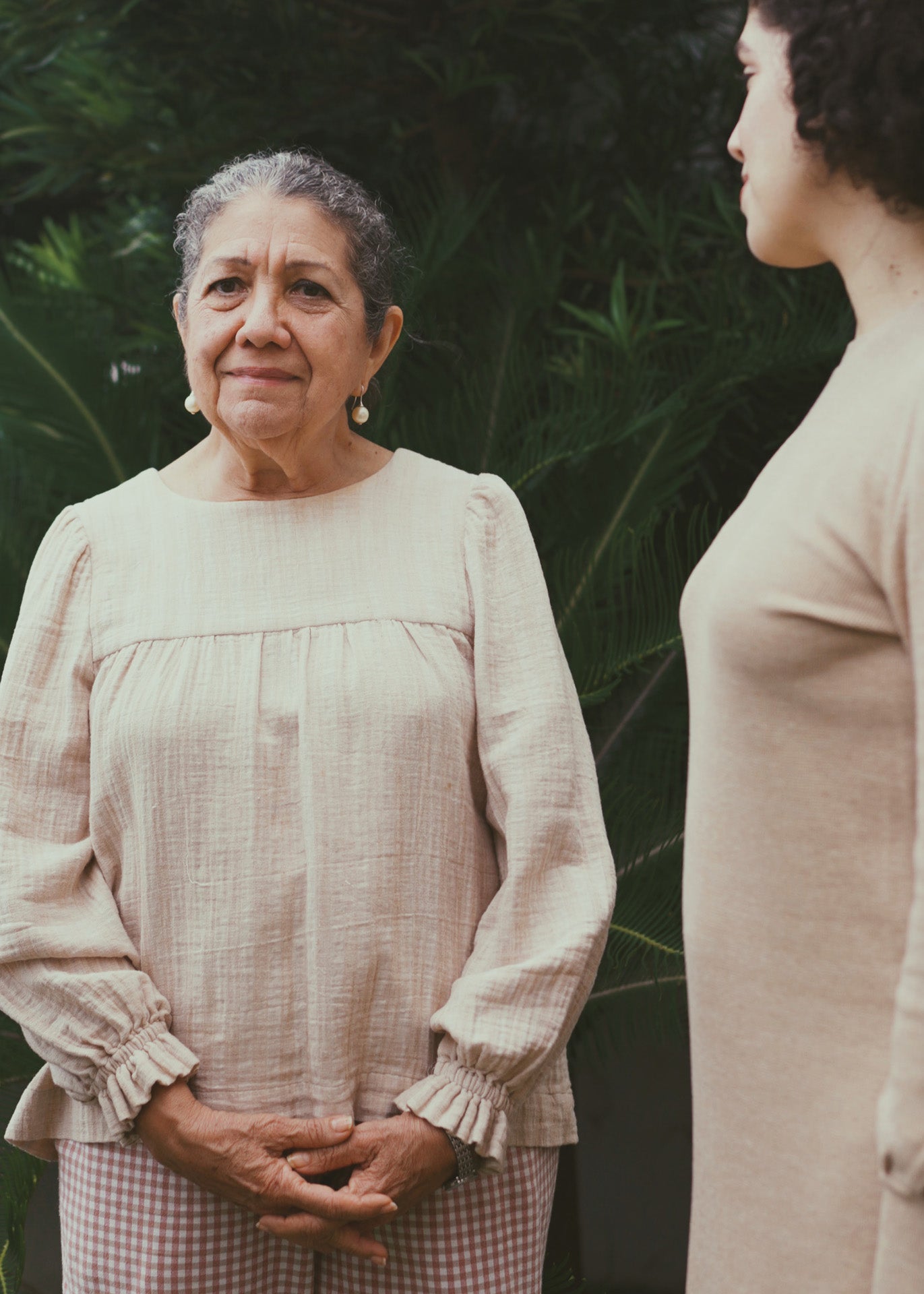 Two women standing outdoors with a natural background. The woman on the left is wearing a blouse with round neckline, gathering details, and long peasant sleeves. The woman on the right is wearing a light brown dress.
