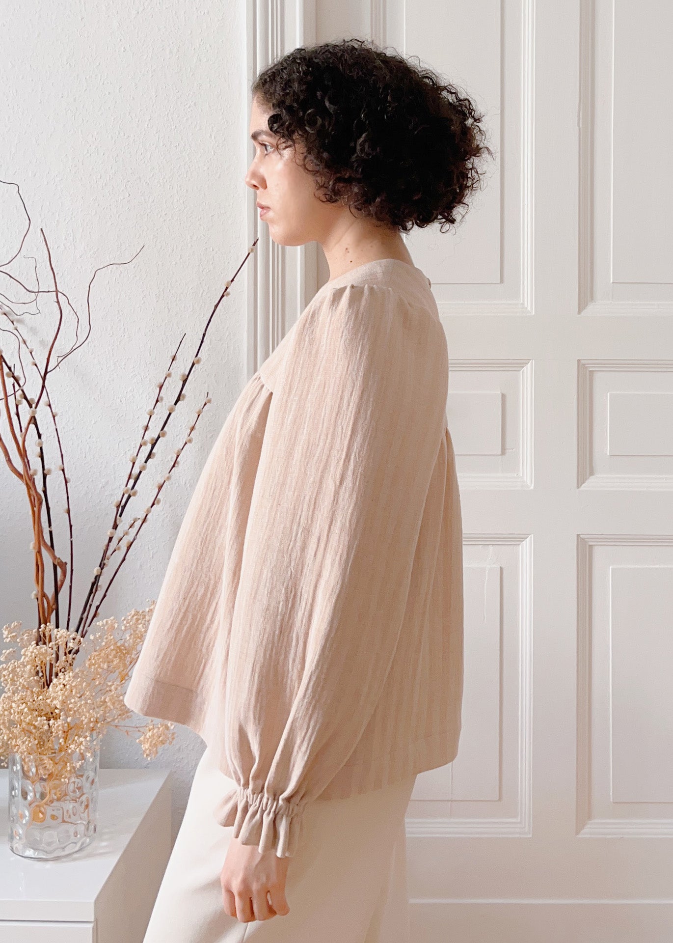 Woman wearing a light brown striped blouse standing in a room with white paneled walls and decorative plants. The blouse has long peasant sleeves with elastic ruffle cuffs.