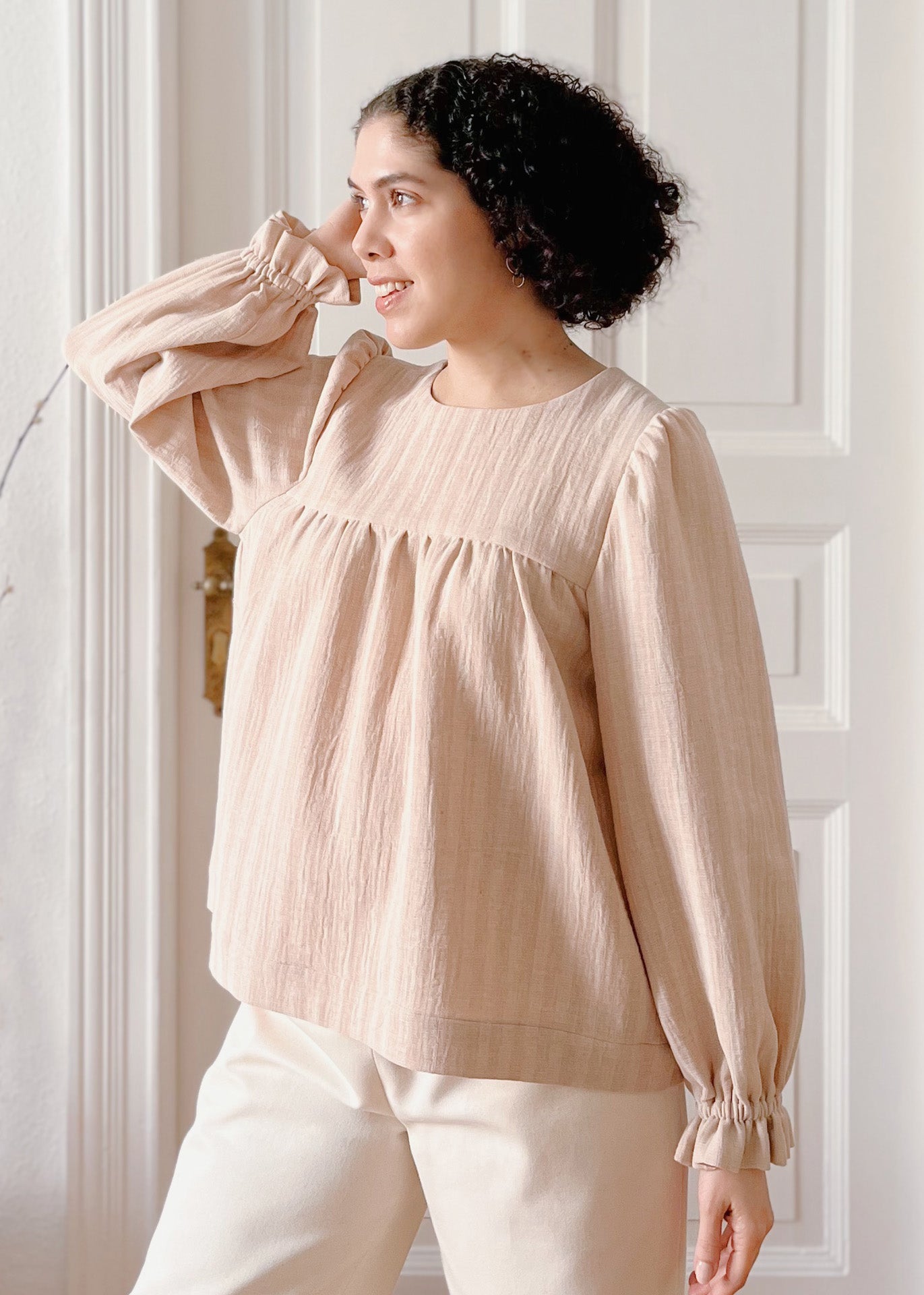 Woman wearing a light brown striped blouse with long sleeves and white pants against a white wall. The blouse has loose fit and classic round neckline.