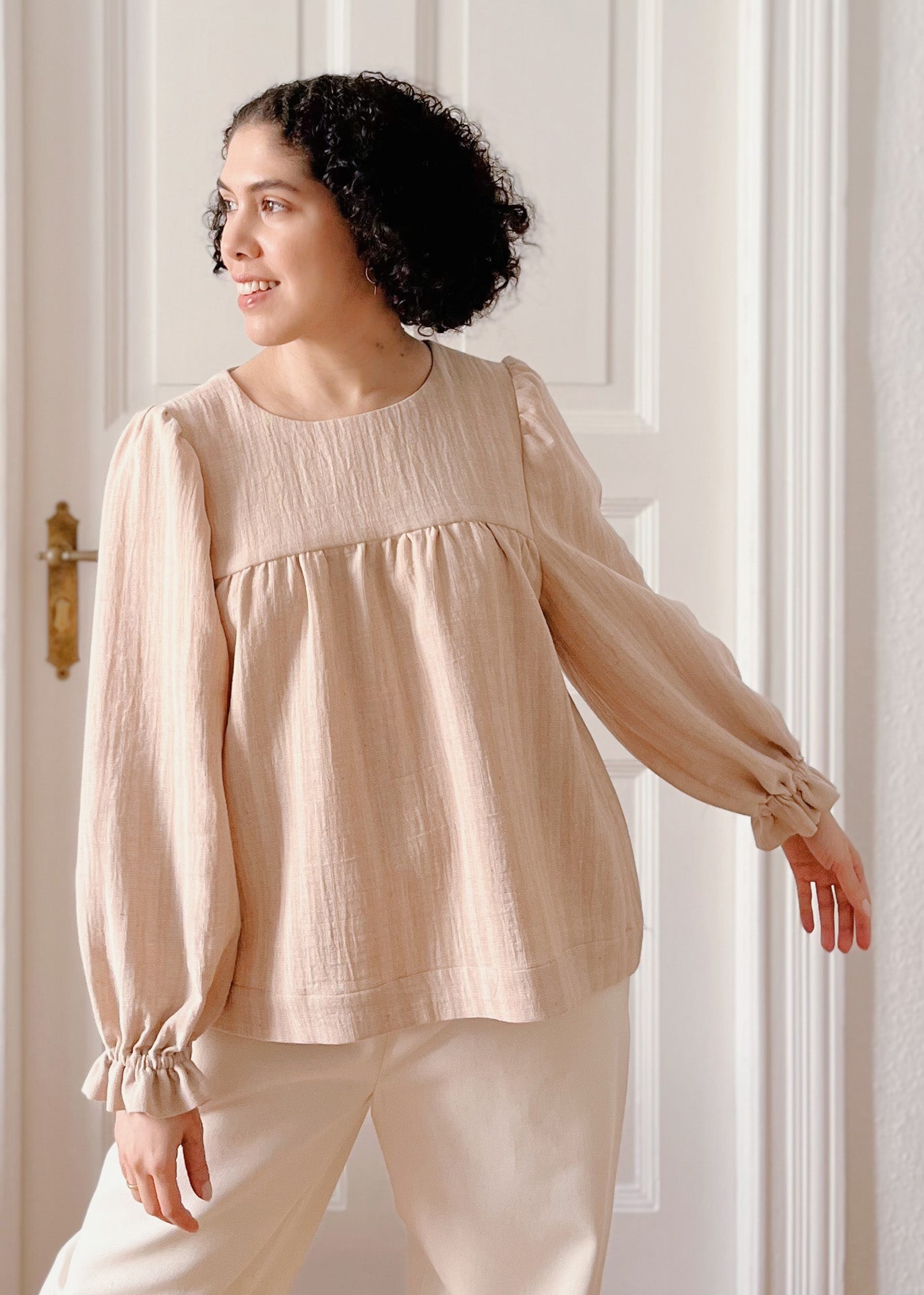 Woman wearing a light brown striped blouse and white pants standing in a softly lit room.