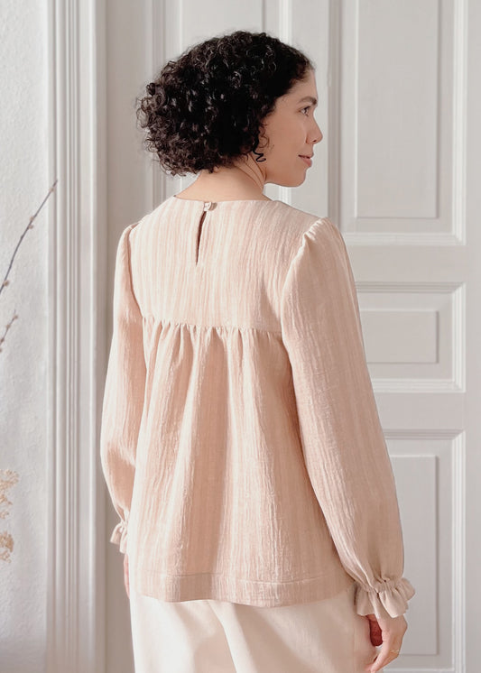 Woman wearing a light brown organic cotton blouse standing against a white paneled wall.
