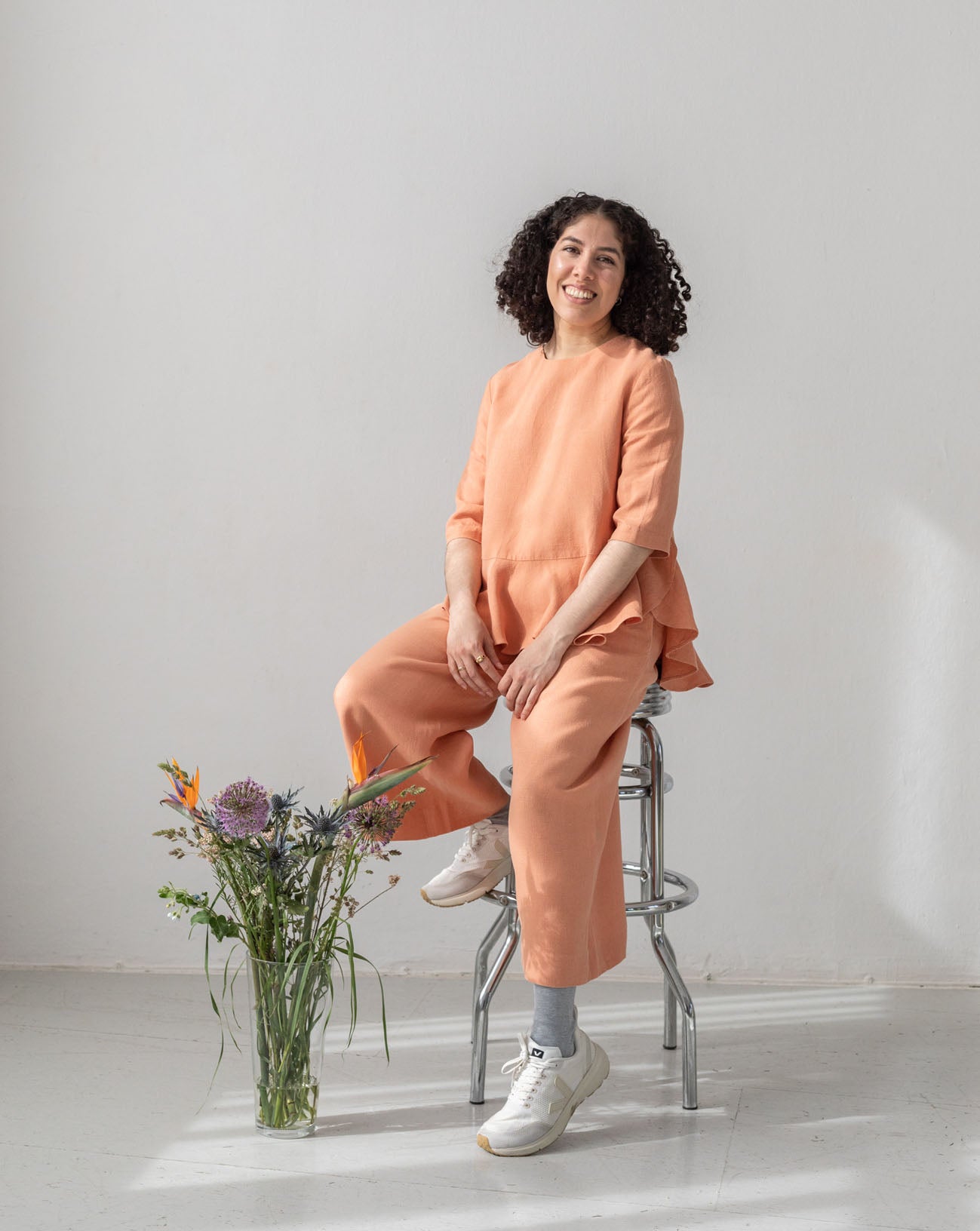 Designer Daniela Salazar sitting on a barstool and smiling. She is wearing eco conscious hemp clothing. The Mara top and Triadic pants, both in peach hemp fabric. Next to her, there is a vase with flowers. Grey background.