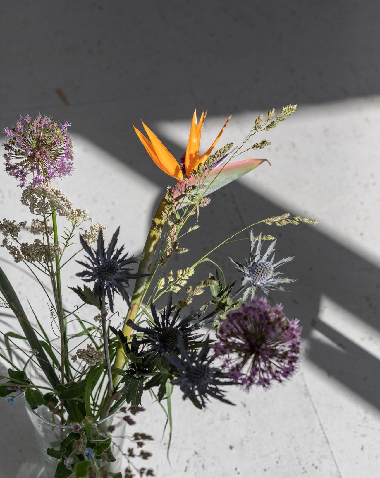 A vase with a mix of flowers in natural colors like green, lavender and orange. Sunlight and shadow in the background.