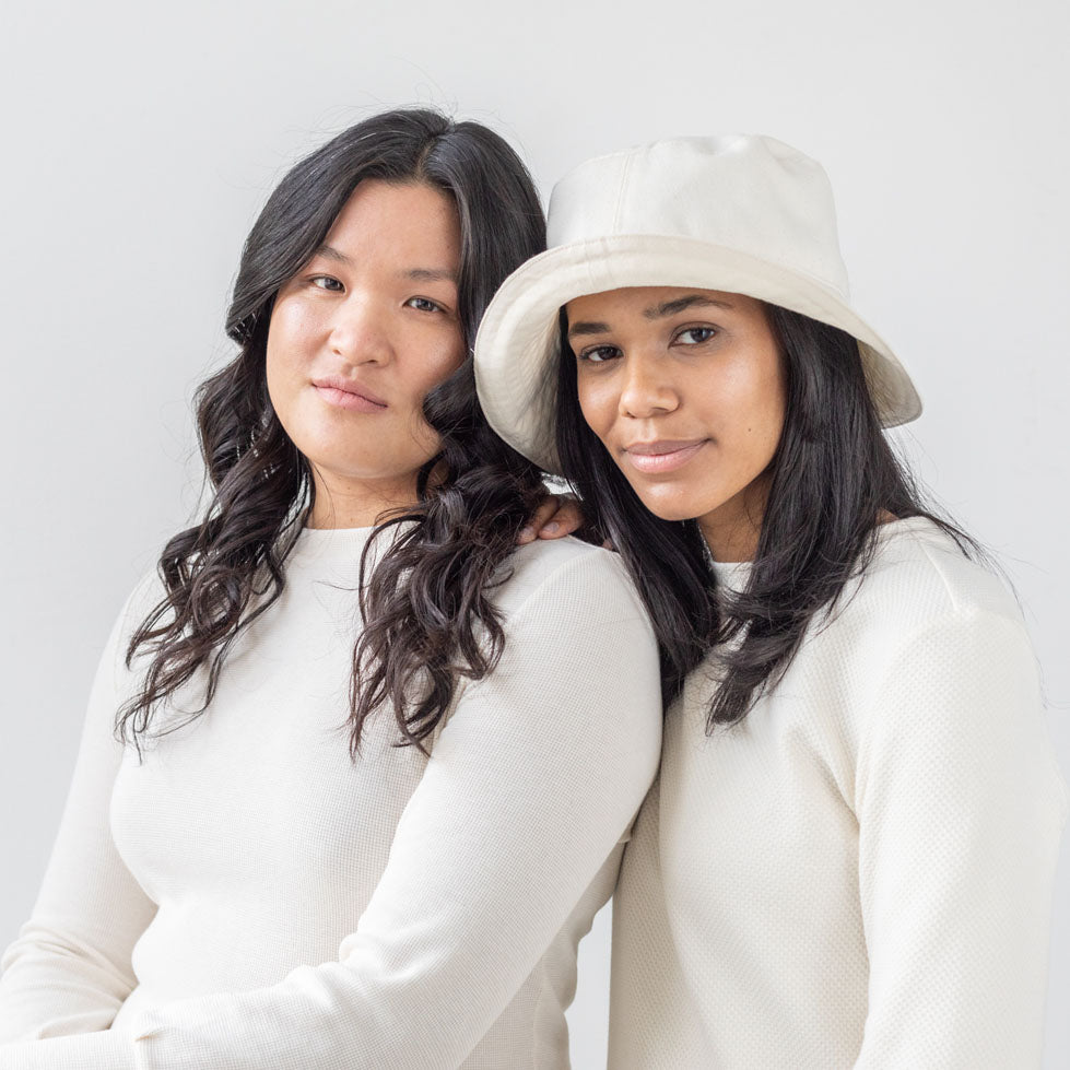 Two women standing head to head and smiling. They are wearing organic cotton dresses with long sleeves. One of them is wearing a white bucket hat.