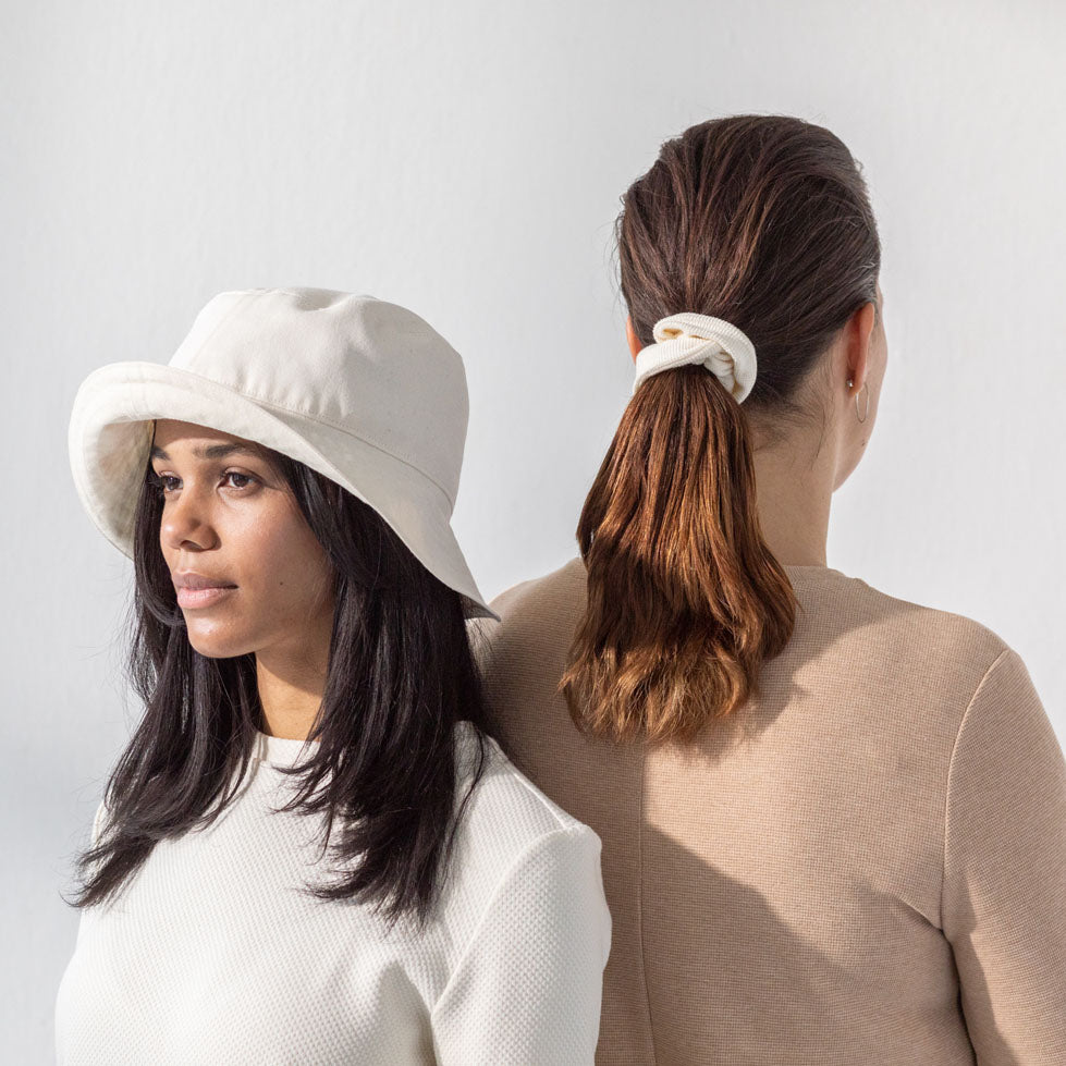 Two women standing back to back. They are wearing organic cotton dresses in natural colors. One woman is facing the background wearing an organic cotton scrunchie on her hair. The other woman is wearing a white bucket hat.