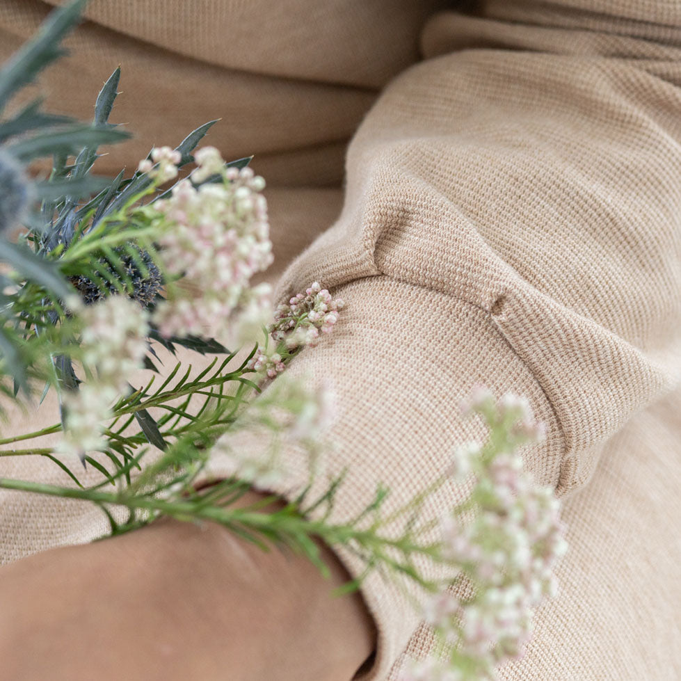 A close up of a woman wearing a dress with sleeve and long cuffs. On her hands she is holding some wild flowers. The organic cotton jersey fabric is textured and light brown.