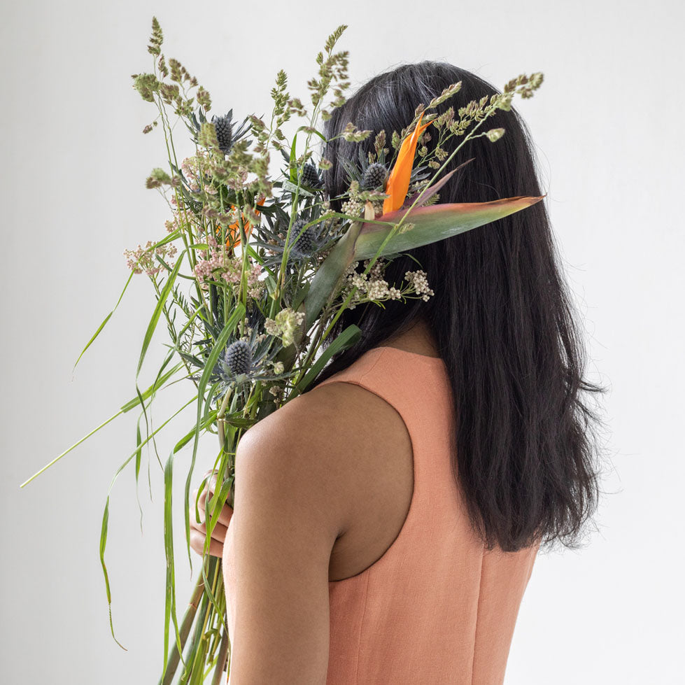 A woman facing a grey background. She is holding a bouquet of wildflowers at shoulder level. The flowers cover her face. She is wearing hemp clothes for women from slow fashion brand Daniela Salazar. The eco friendly dress is made in peach hemp fabric