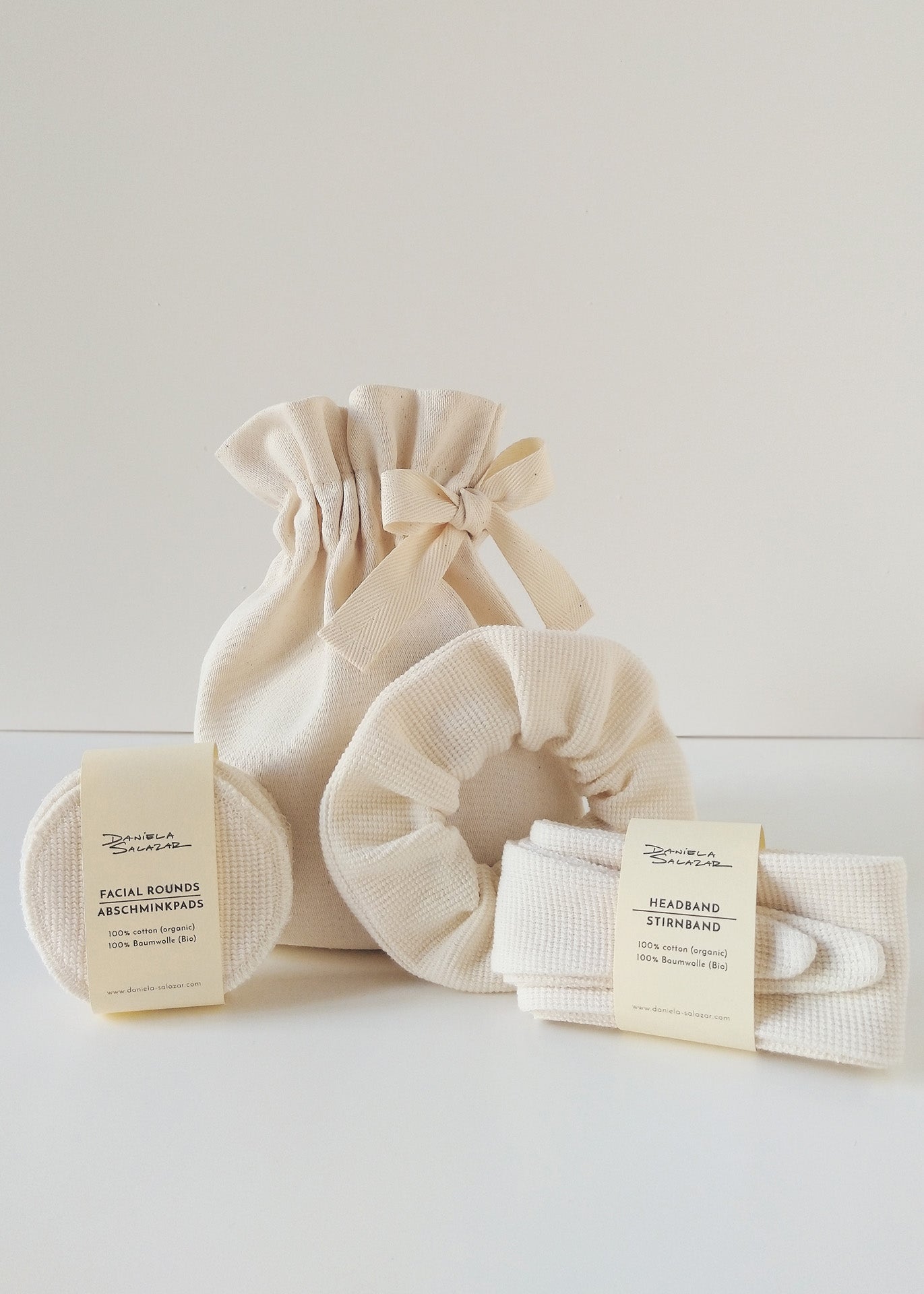A set of ivory-colored home spa accessories including a headband, reusable face pads and a scrunchie, packaged in an ivory cotton gift-ready pouch.