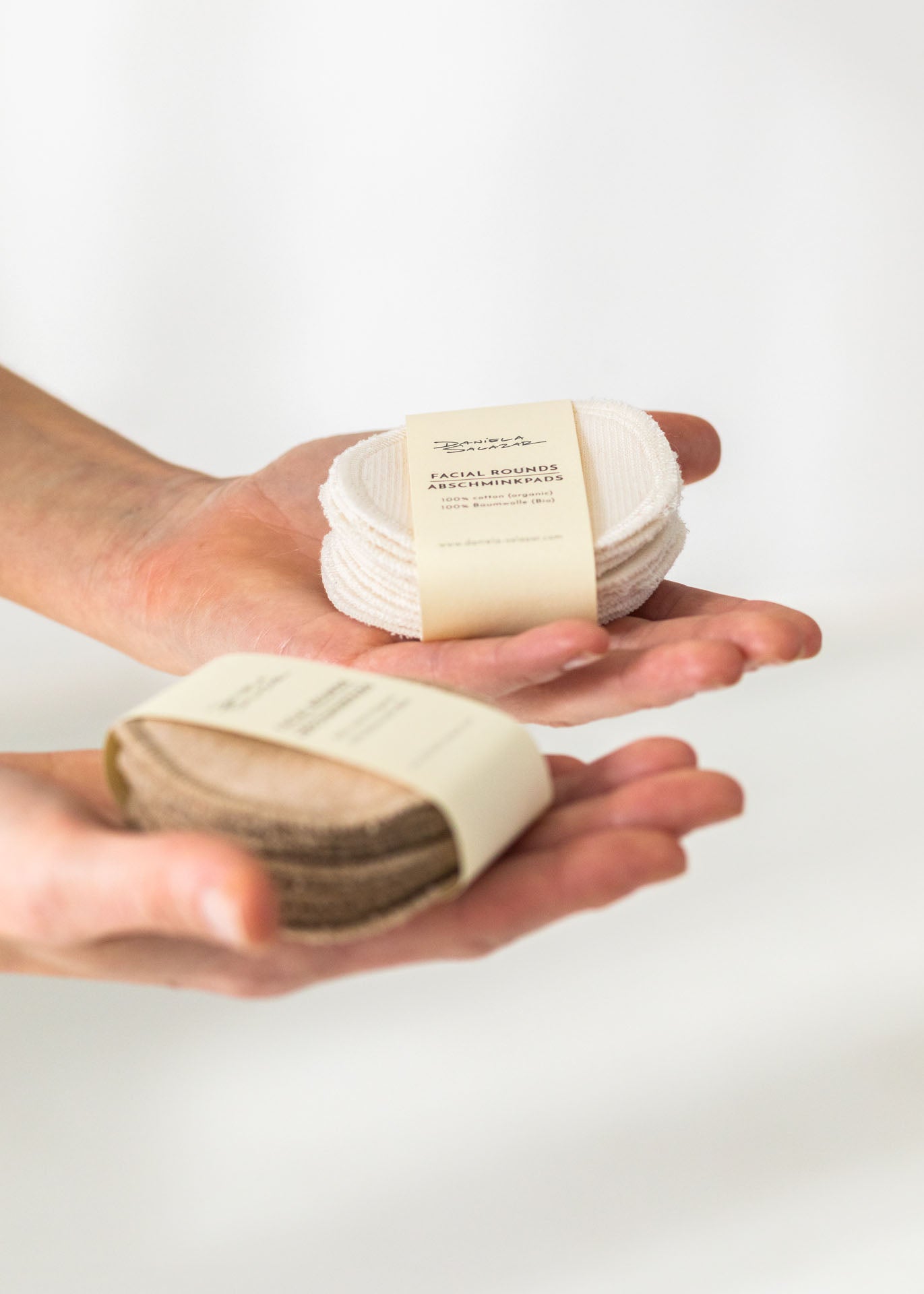 A close up of two hands holding organic cotton face pads, one pack in ivory and one in brown. These cotton facial pads are a zero-waste design.