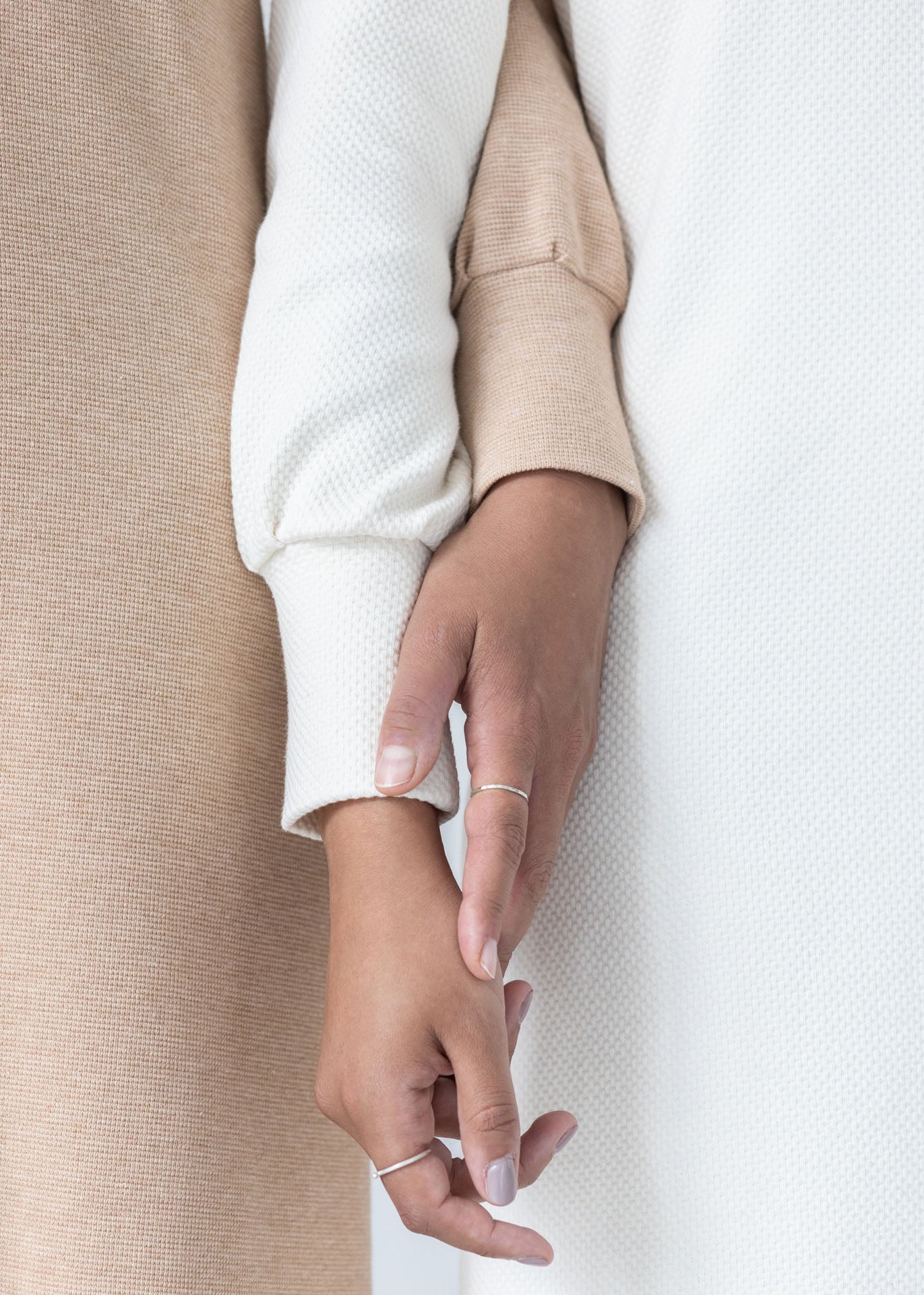 Detail of two hands touching. One woman is wearing a white dress, the other woman is wearing a brown dress. Focus on the textures of the organic cotton jersey fabric.