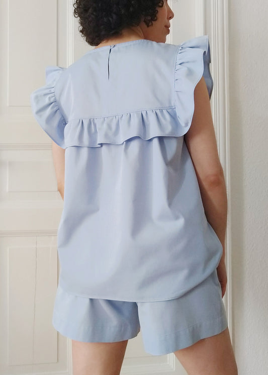 Woman wearing the Tana Blouse in sky blue, showing the back side with ruffles bib, keyhole and button closure.