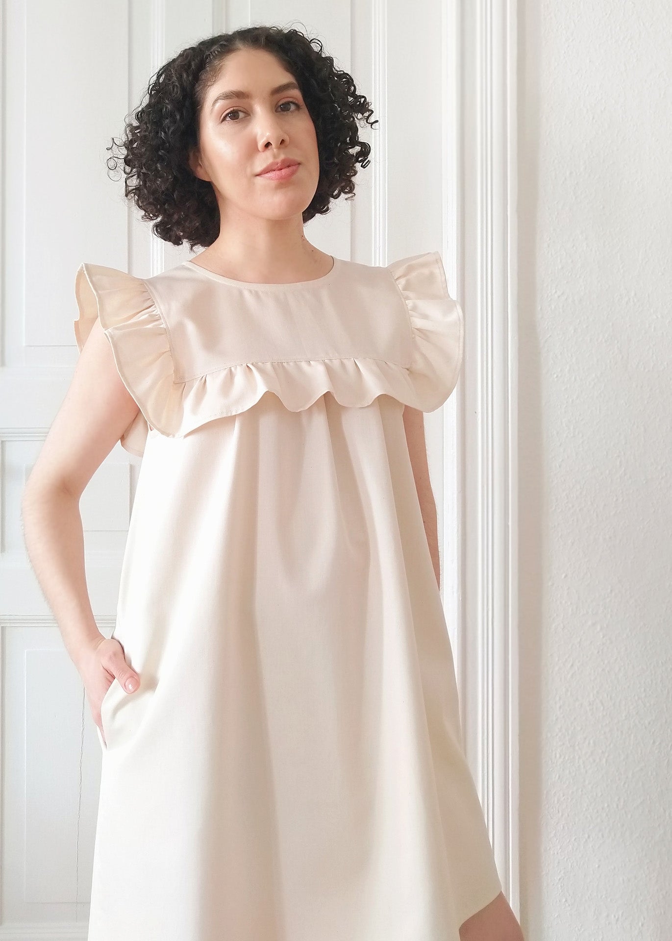 Woman wearing a Hana Ruffle Mini Dress in Ecru organic cotton standing against a white paneled wall.