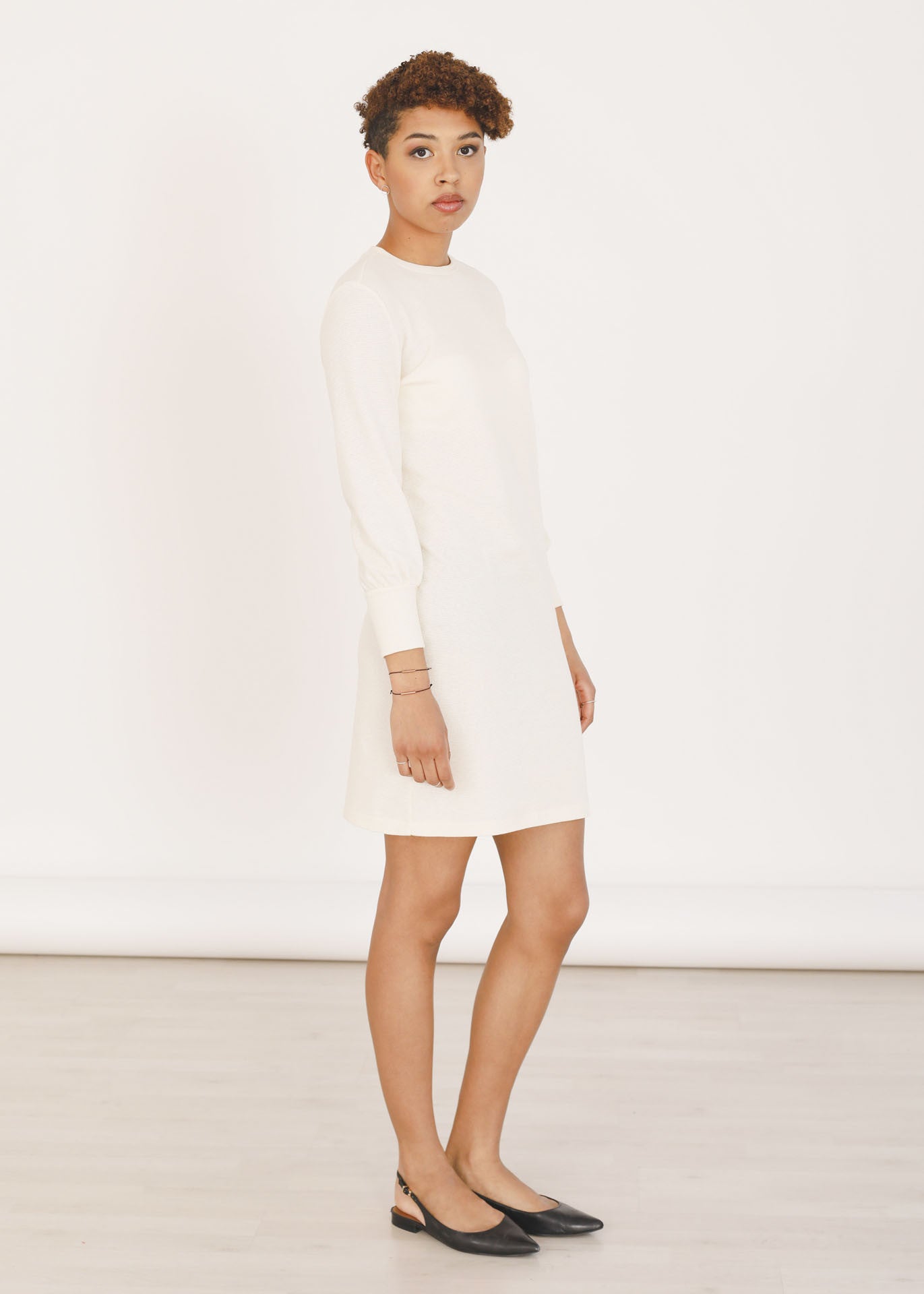 A woman wearing an Ivory long-sleeved sweater dress with a crew neckline, made of organic cotton, standing in front of a white background.