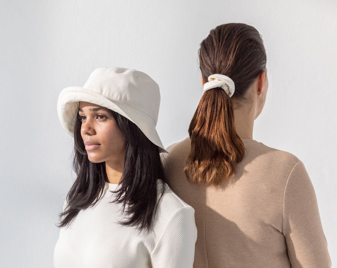 Two women standing back to back. They are wearing organic cotton dresses. One woman is facing the background wearing an organic cotton scrunchie on her hair. The other woman is wearing a white bucket hat.