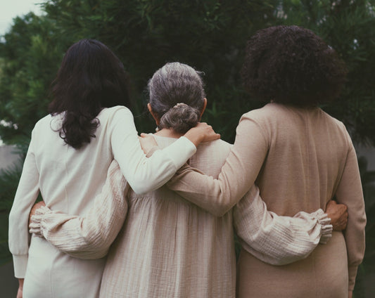 Three women standing together facing back. They are holding each other and wearing muted colors clothing. There is greenery in the background.