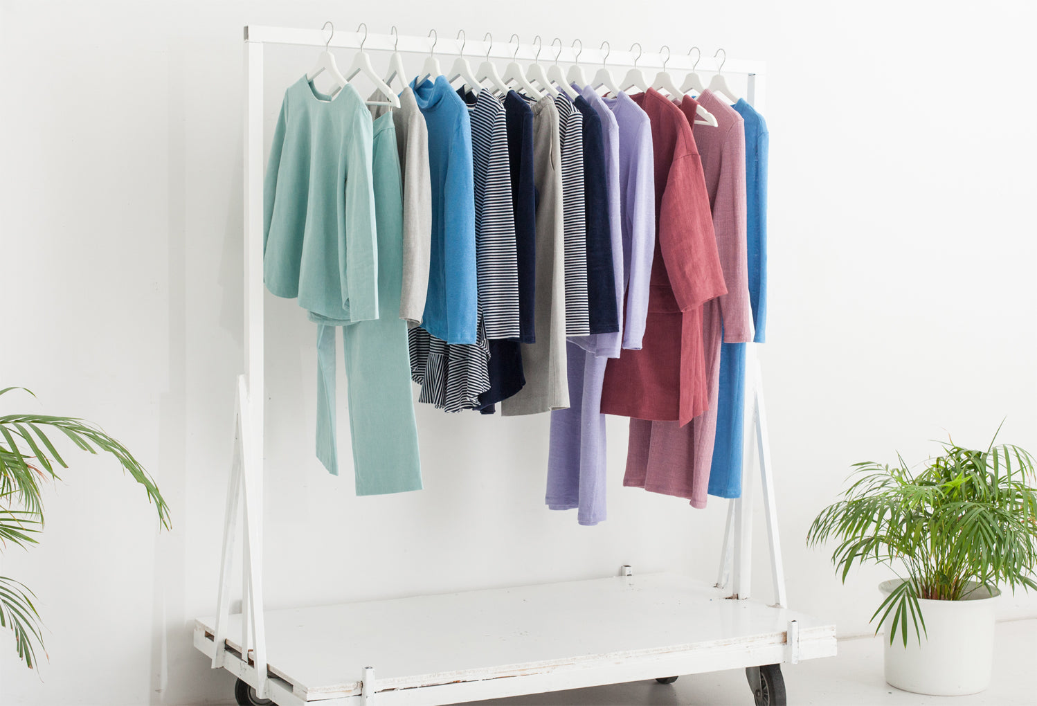 Clothing rack with various eco friendly tops, pants and dresses hanging on it against a white wall and plants on both sides. 