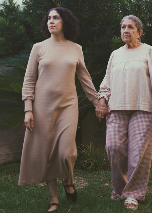 Two women holding hands outdoors with a natural background. The woman on the left is wearing a midi brown dress with long sleeves.