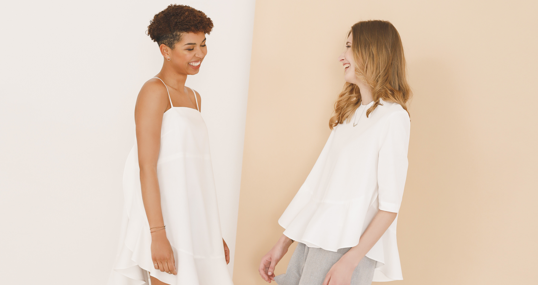 Two women against a white and beige background. They are facing each other and smiling. The model on the left is wearing a white short dress and the model on the right is wearing a white top and grey shorts.