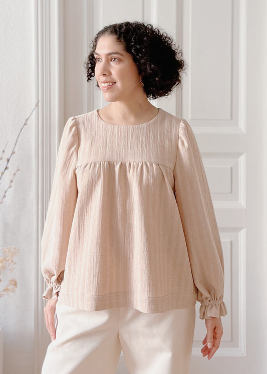 Woman wearing a light brown striped blouse and white pants against a white paneled wall.