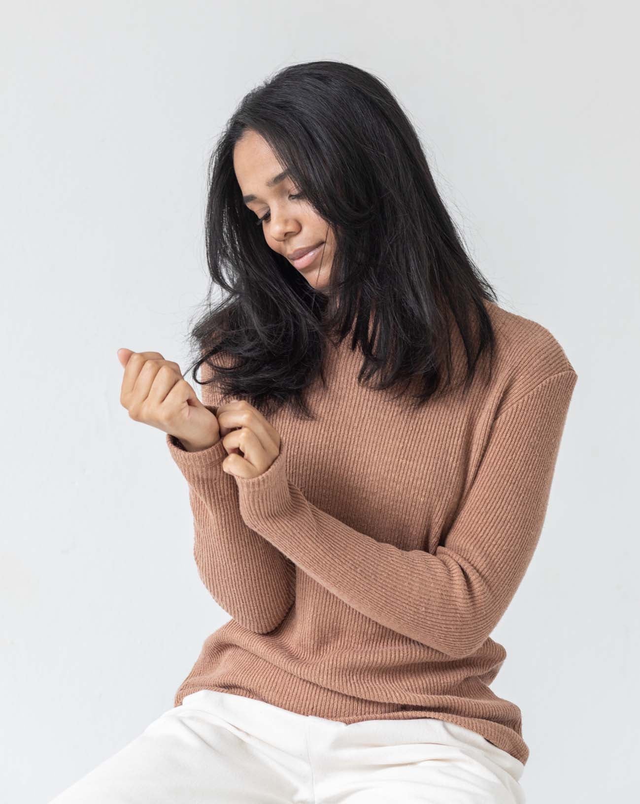 Woman wearing a brown PARK turtleneck sweater and white TRIADIC pants against a white wall. She is touching and looking at her sleeve.