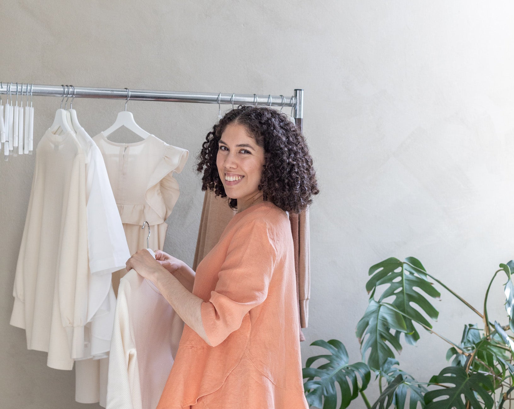 The slow fashion brand designer Daniela Salazar is holding a Tana dress in ecru organic cotton. In the background there's a clothing rack with her designs and some greenery. She's wearing the Mara top and Triadic pants, both in Coral hemp fabric. She's looking and smiling.