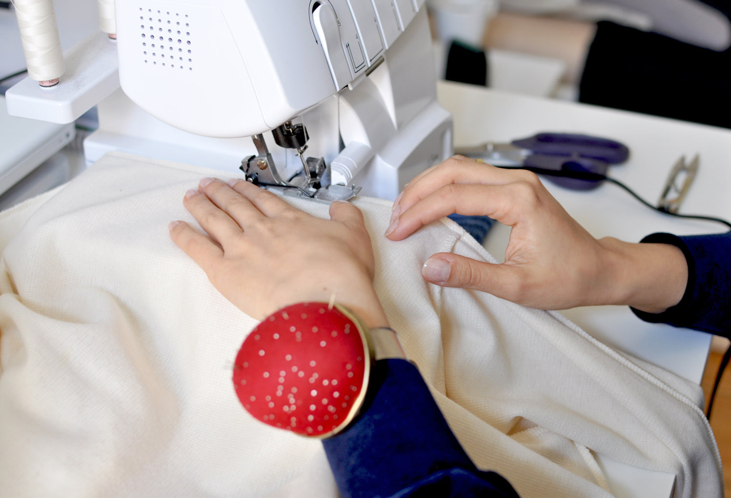 Close up of two hands and a sewing machine during the process of sewing an organic cotton dress. There are scissors on the table. On the left arm there is a pin cushion.