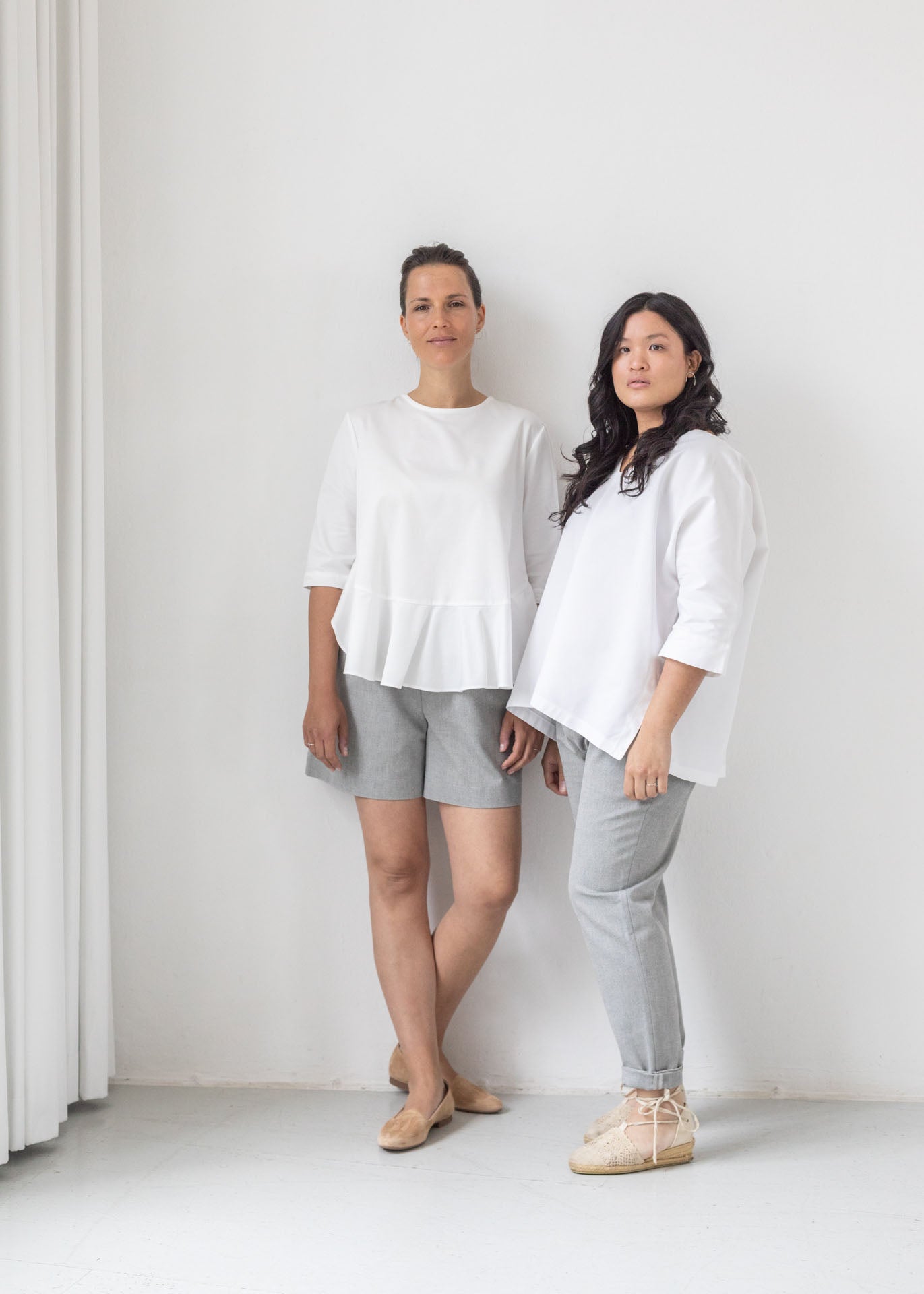 Two women stading against a white wall. They are both wearing white organic cotton tops and grey bottoms. 